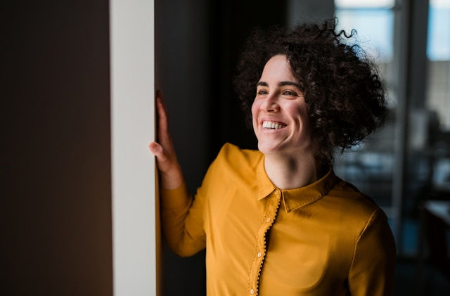 A smiling person with curly hair leans on a wall in an office setting, wearing a yellow shirt. Sunlight filters through large windows.