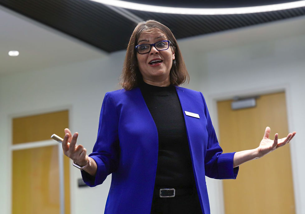 Woman in a blue blazer speaks animatedly with open hands in a modern office hallway, holding a small remote, smiling confidently