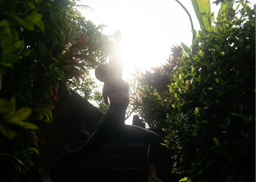 Silhouetted person in a yoga pose on steps, surrounded by lush greenery. Bright sunlight creates a serene, peaceful atmosphere.