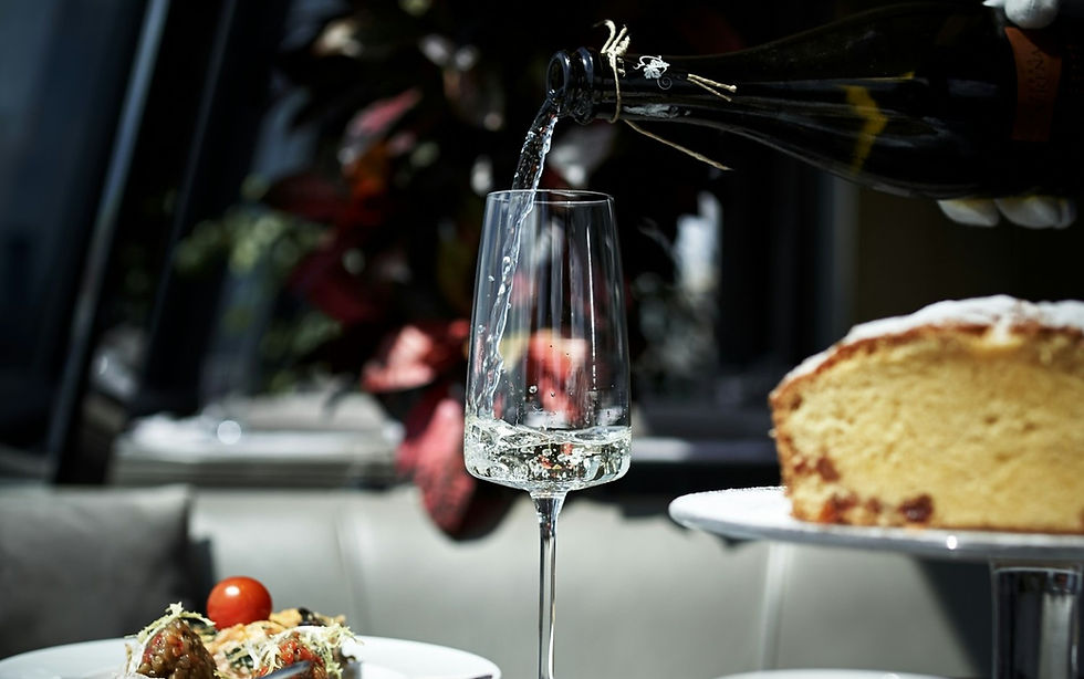 Champagne pours into a flute beside a slice of cake in a dim restaurant, with plated food in the foreground.