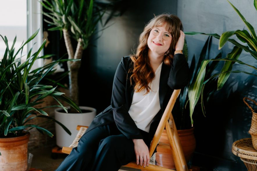 A photo of Katie with long red hair and glasses sits barefoot on a white office chair in a cozy, art-filled home office, smiling confidently beside a dual-monitor setup.