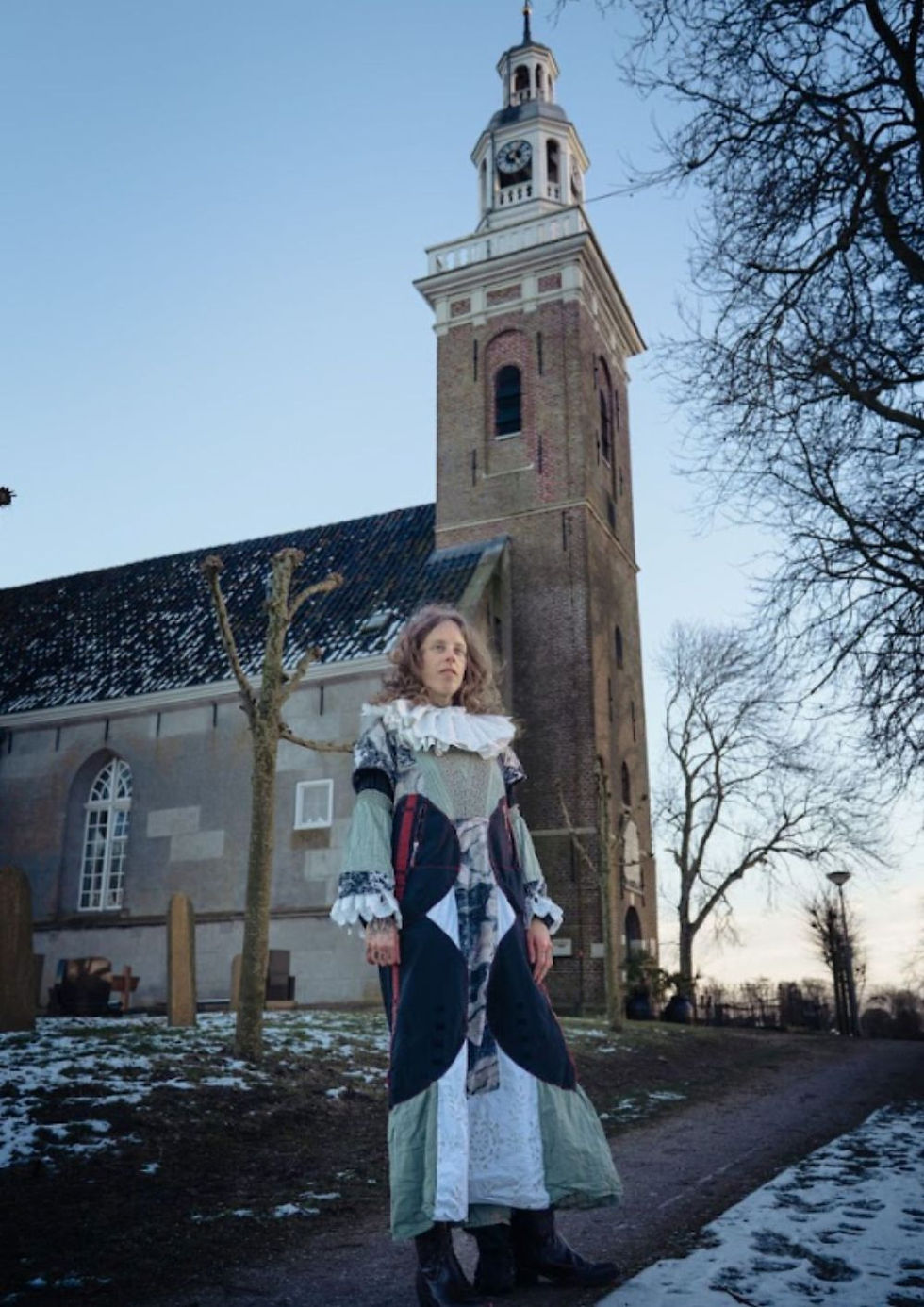 The photo shows a woman in a vintage-style dress standing in front of a church tower, with a snowy path and trees around her.