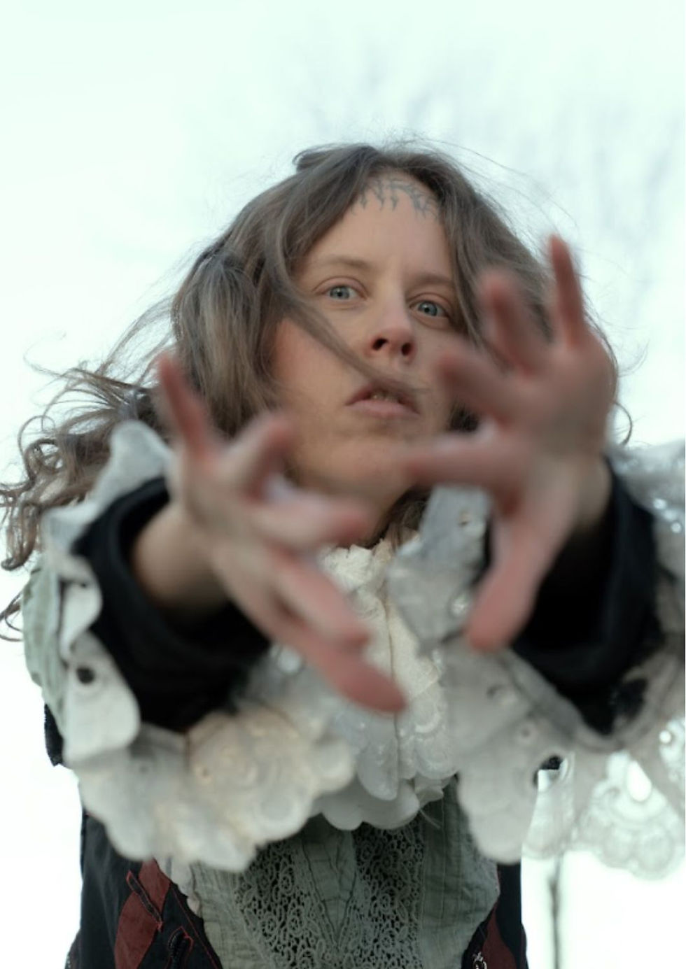 Person with long hair and lace sleeves reaches towards the camera with an intense expression. Pale sky in the background.