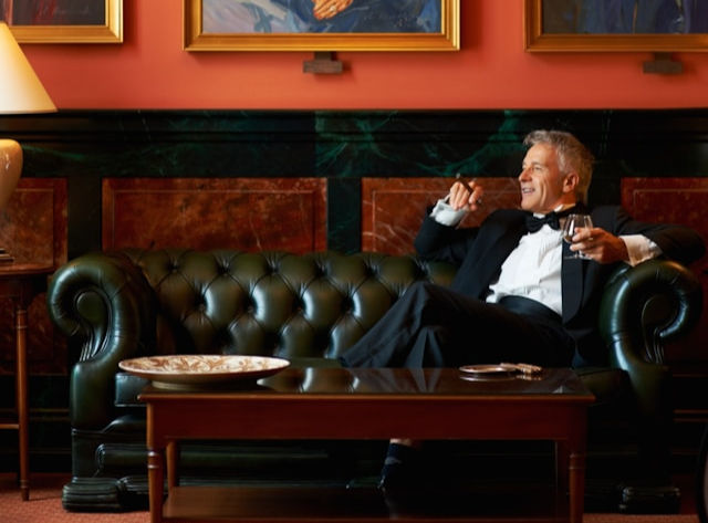 Man in tuxedo relaxes on a green leather sofa, holding a glass. Elegant room with paintings, warm lighting, and a sophisticated atmosphere.