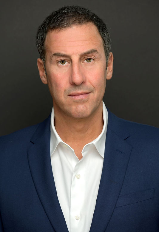 Man in a blue suit and white shirt, neutral expression, against a plain dark background.