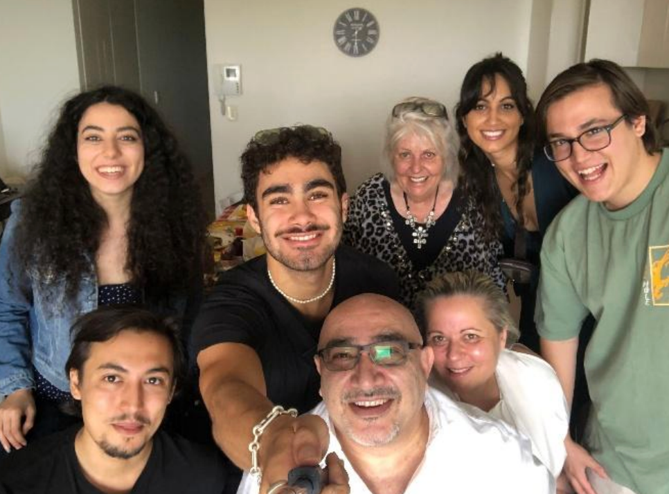 Smiling group selfie of eight people indoors, with an older woman and man in front, in a cozy room with wall clock.