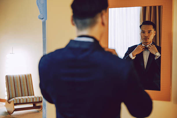 Man in a suit adjusts bow tie in front of a mirror, reflecting a calm expression. Background features a modern chair and soft lighting.