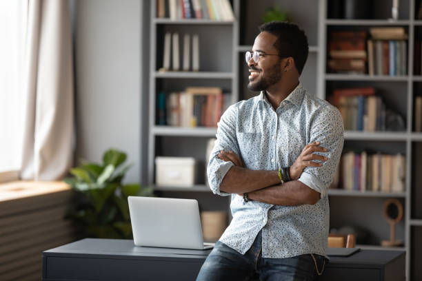 A smiling man with glasses stands with his arms crossed in front of a desk with a laptop, in a room with bookshelves.