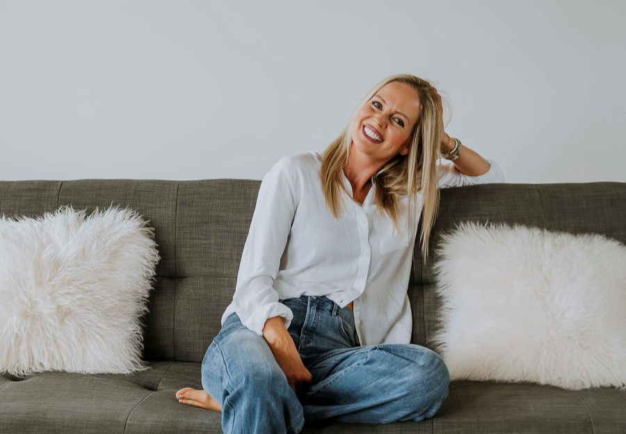 Woman in white blouse and jeans smiles, sitting on a gray sofa with fluffy white pillows. Bright, casual setting with a relaxed mood.