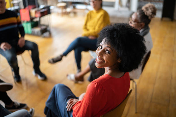 Smiling woman in a red shirt sits in a circle with others on wooden floor. Casual, friendly setting. Bright and warm atmosphere.