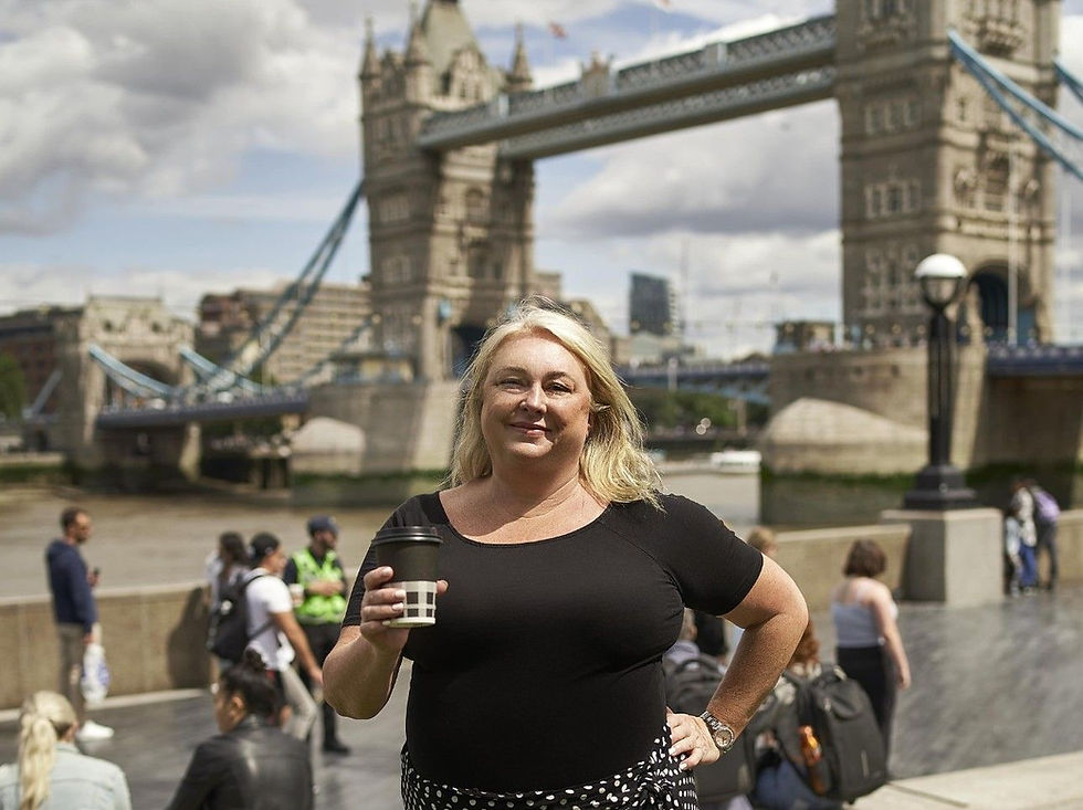 A woman in a black top holds a coffee cup near Tower Bridge, London. It's sunny with people in the background, creating a lively mood.