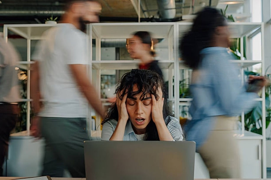 Overwhelmed woman sitting at a laptop with her hands on her head while coworkers move quickly around her in a busy office.