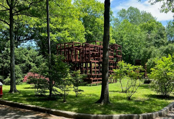 Sunlit park with a rust-red geometric metal sculpture among lush green trees and grass.