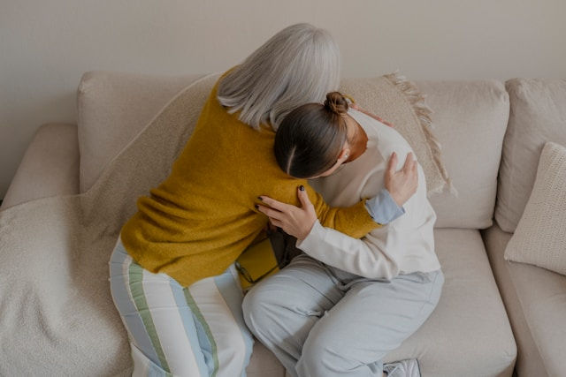Two women sit on a beige couch in a warm embrace, one comforting the other in a quiet, intimate home setting.