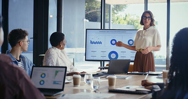 Woman presents financial analytics charts on a screen during a meeting, with coworkers seated around a conference table in a bright office.