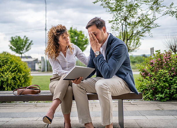 Woman comforts distressed man on bench with tablet. Overcast day, trees in background. Brown bag on bench, professional attire.