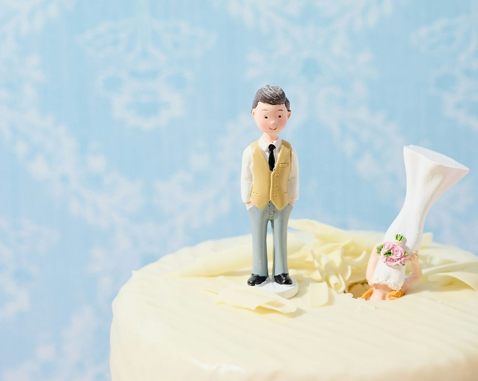 Cake top with a standing groom figurine and a bride figurine upside down in the frosting, against a light blue patterned background. Playful mood.