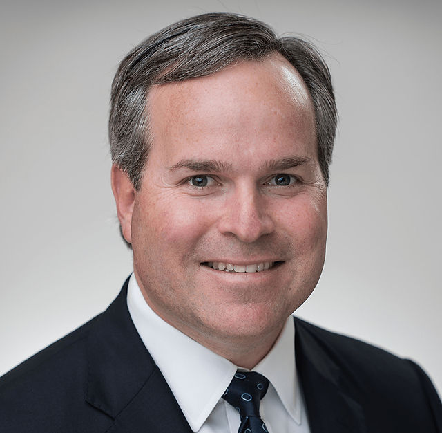Man in a suit with a white shirt and navy tie, smiling. Gray background, professional headshot. Mood is confident and friendly.
