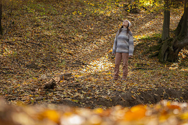 Woman in a gray sweater stands in a leaf-covered autumn forest, looking up among golden trees.