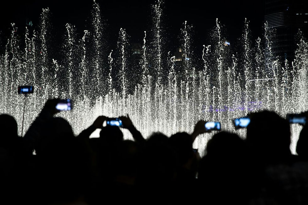 The photo captures a nighttime fountain show with water jets illuminated against the dark sky. Silhouetted spectators in the foreground hold up smartphones, recording or photographing the display.
