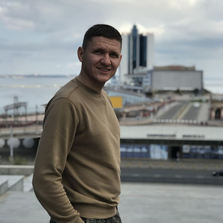 Smiling man in a tan sweater stands by a waterfront cityscape under cloudy skies, with tall buildings blurred behind him.