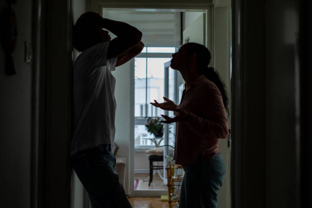 Two people argue in a dimly lit hallway. The man leans against the wall, hands on head; the woman gestures emotionally. A window view behind.