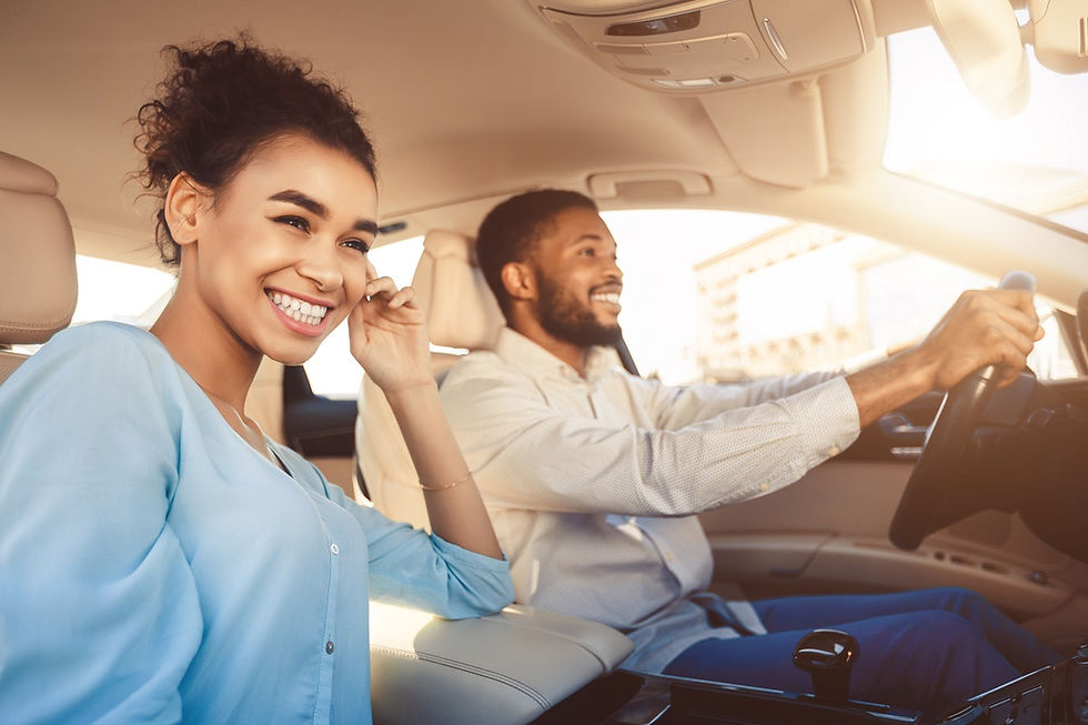 A smiling woman and man drive in a car during daytime. The interior is beige and well-lit, creating a cheerful and relaxed atmosphere.