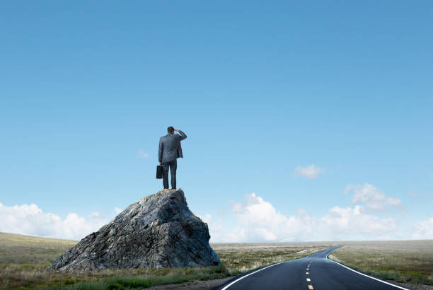 Man in a suit, holding a briefcase, stands on a rock, looking at the horizon. Empty road stretches forward. Clear sky, sense of anticipation.