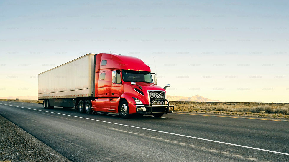 Red semi truck hauling a white trailer drives along a desert highway under a clear sky and distant mountains.
