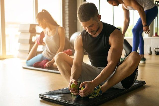 Man stretching on a mat in a bright yoga studio, with two women exercising in the background under warm sunlight.
