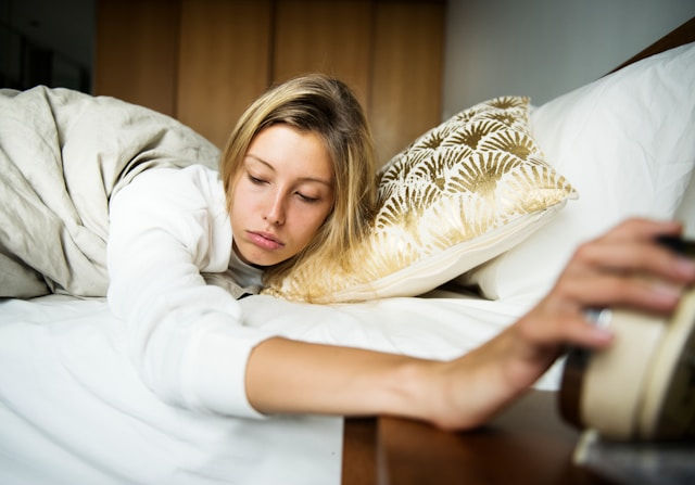 Woman in bed reaching for alarm clock, looking tired. Beige bedspread, gold-patterned pillow. Bedroom setting with a relaxed mood.