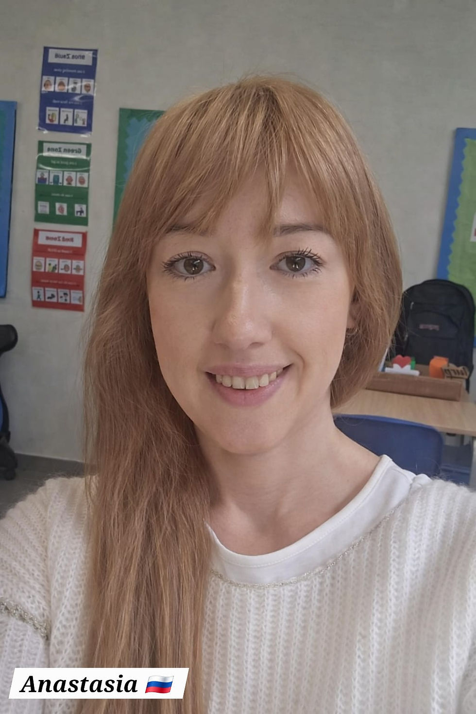 Woman smiling in a classroom with educational posters. Brown hair, white sweater. Name tag reads Anastasia with a Russian flag.