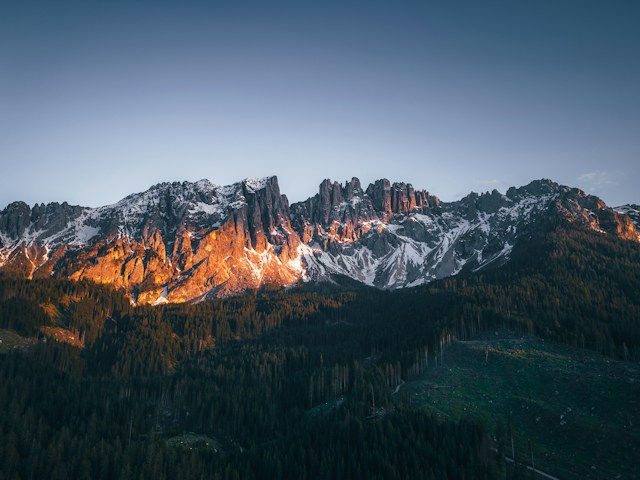 Snow-capped mountain range glowing orange at sunset above a dark pine forest under a clear blue sky