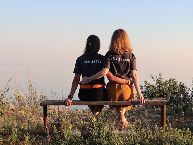 Two women sit closely on a bench with arms around each other, wearing matching "SISTERHOOD" shirts, overlooking a peaceful, natural landscape.