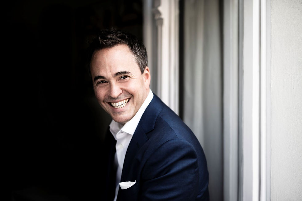 Garrett Keast in a dark suit stands by a window, softly lit background. He exudes happiness with a bright, genuine smile.