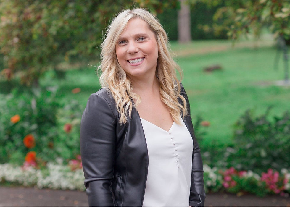 Smiling blonde woman in a black jacket and white top stands in a green garden with flowers.