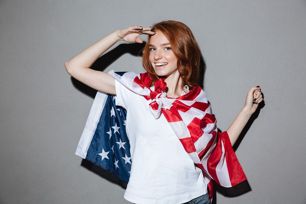 Smiling woman with red hair draped in a US flag, saluting. She's in a white shirt against a gray background, conveying a patriotic mood.