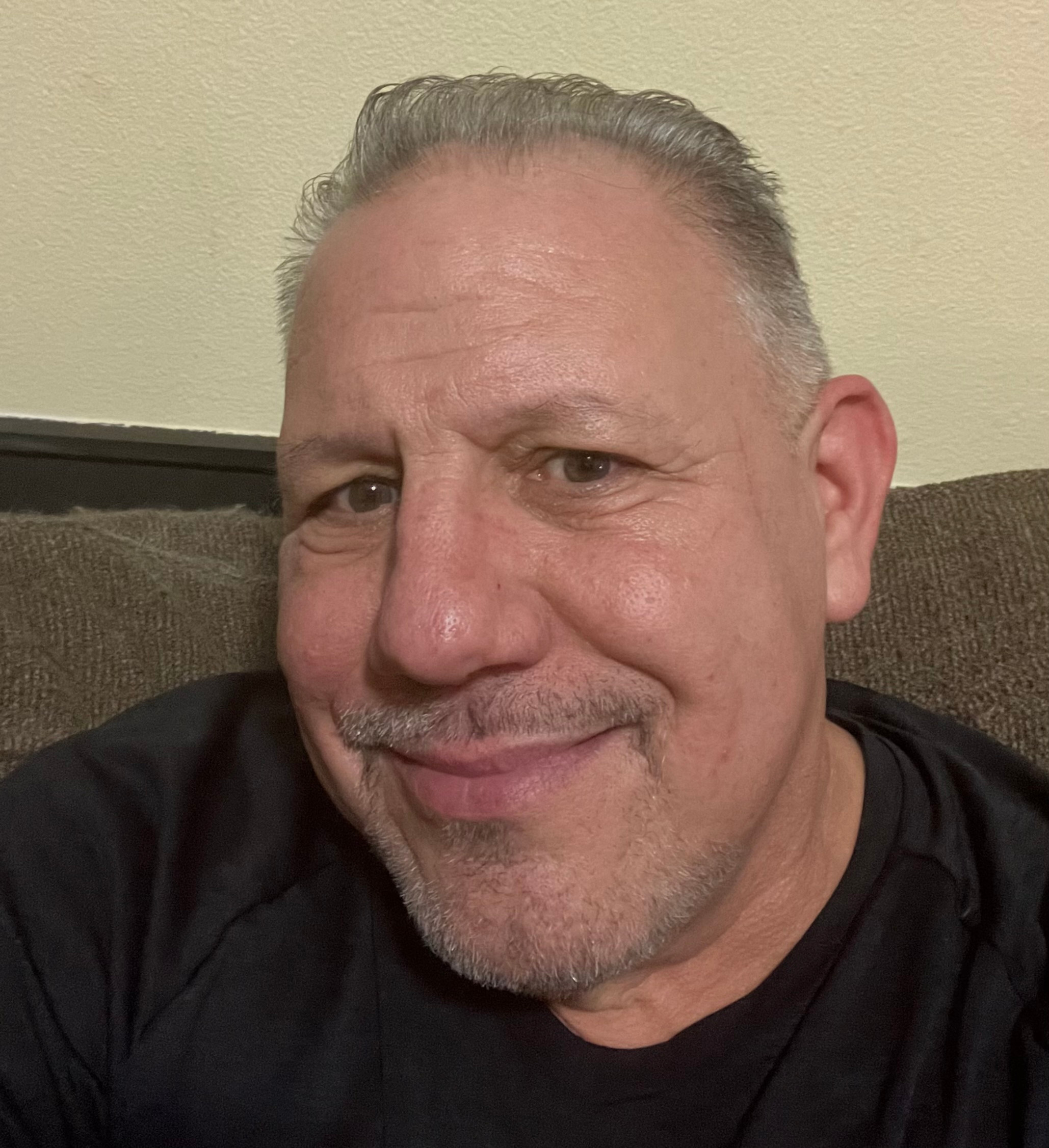 Close-up selfie of a smiling middle-aged man with gray hair and goatee, sitting on a couch against a beige wall.