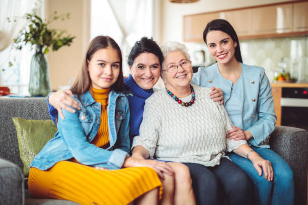 Four women of different ages sit together on a sofa, smiling warmly. They're in a cozy living room with soft lighting and plants.