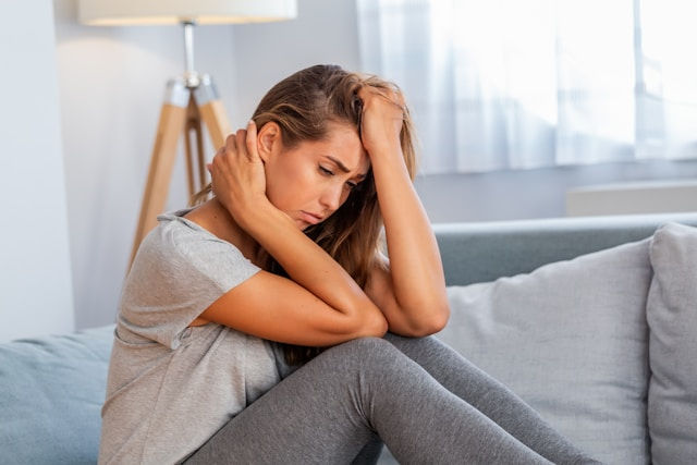 A woman sitting on a couch, appearing distressed or deep in thought, with her hands holding her neck and head.