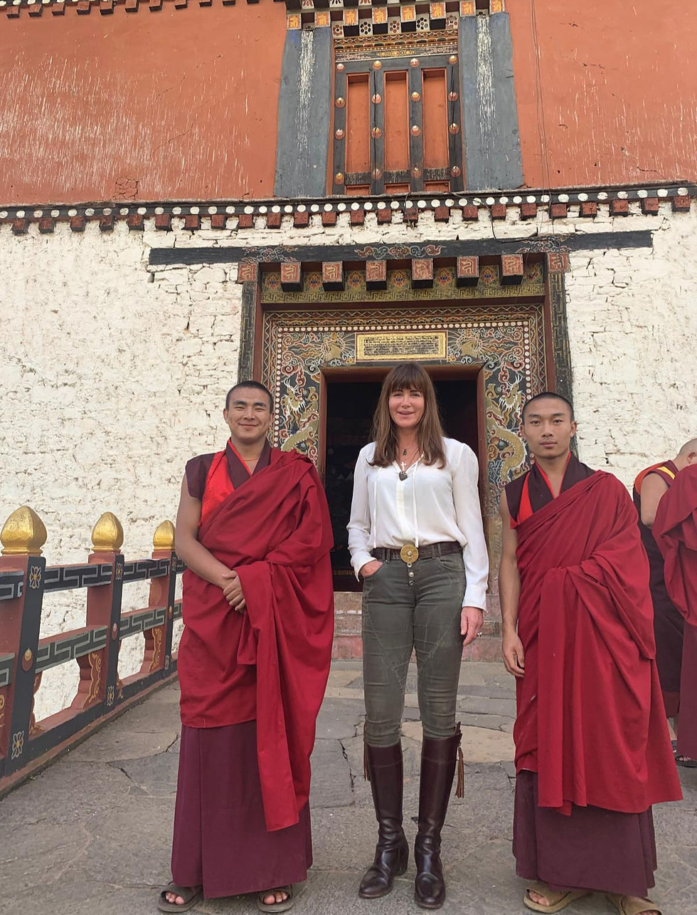 Woman poses with two monks outside an ornate Tibetan temple, with red, white and gold painted walls and doorway.