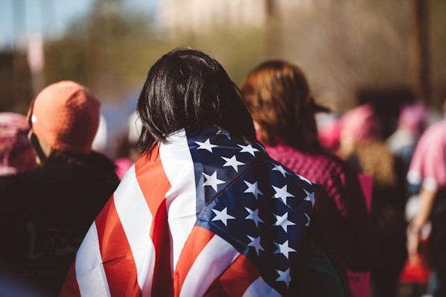 A person is seen from behind wearing an American flag draped over their shoulders in a crowd during a public event or gathering.