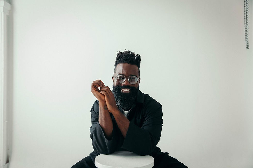 Smiling man with glasses and beard, wearing black, leans on a white stool against a plain white background. Relaxed and cheerful mood.