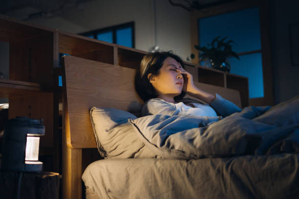 Woman lying in bed at night, hand to forehead, looking tired or in pain, with a warm lamp and blue window behind her.