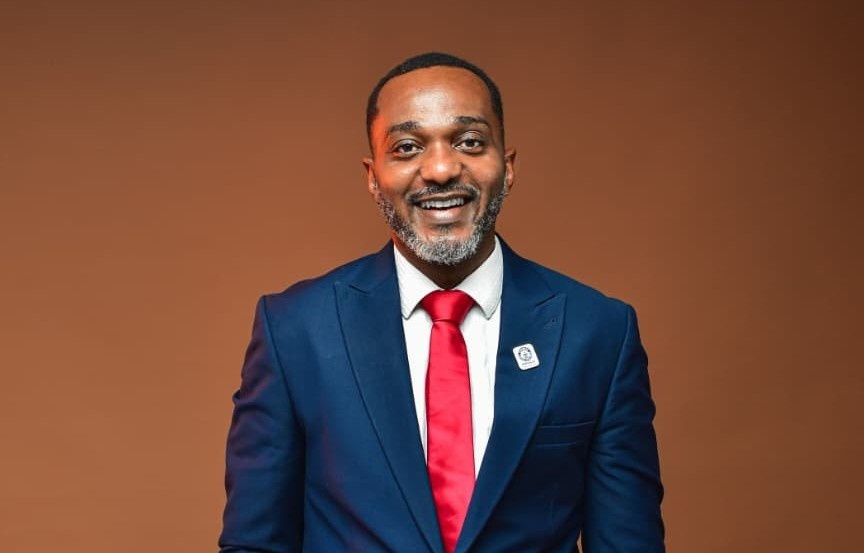 Smiling man in blue suit with red tie stands against a brown background, wearing a pin. He conveys a professional and positive demeanor.