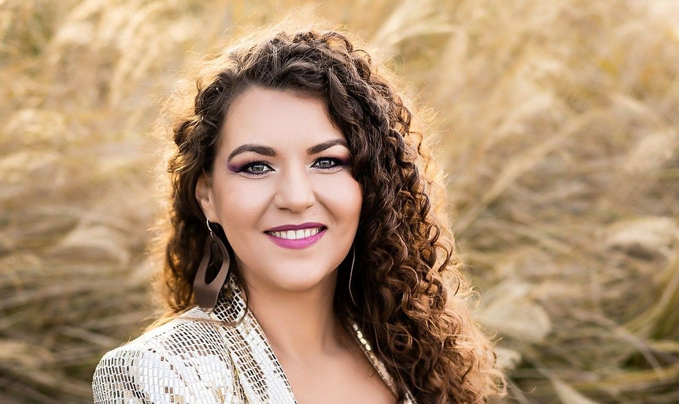 Smiling woman with curly hair in a sunlit field, wearing a light patterned jacket and earrings, in a warm outdoor portrait