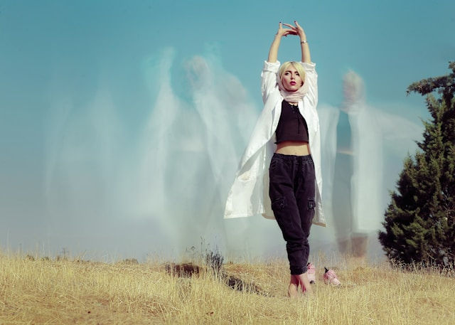 A woman poses with arms raised in a dry, grassy field. She wears a white coat and dark pants. Blurred ghostly figures appear behind her.