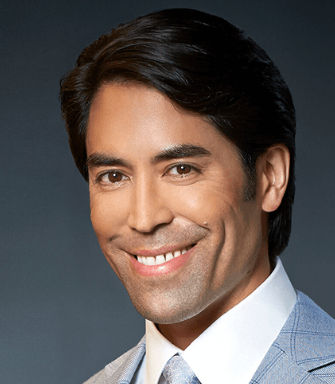 Smiling man in a light blue plaid suit and tie against a dark background. Emotion is cheerful and professional. No text visible.