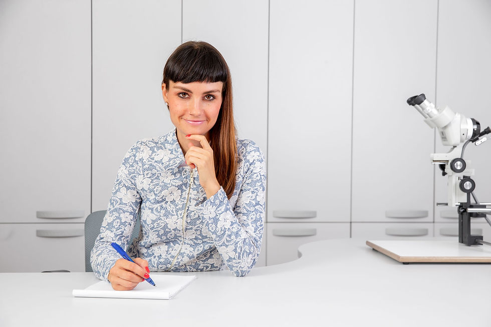 Woman in a floral top writes in a notebook at a desk beside a microscope, smiling in a bright lab office.