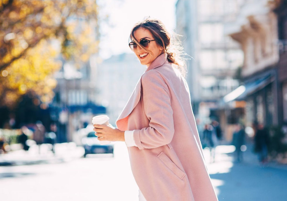 Woman in a pink coat, smiling with sunglasses and a coffee cup on a sunny street. Blurred urban background, blue sky, autumn trees.